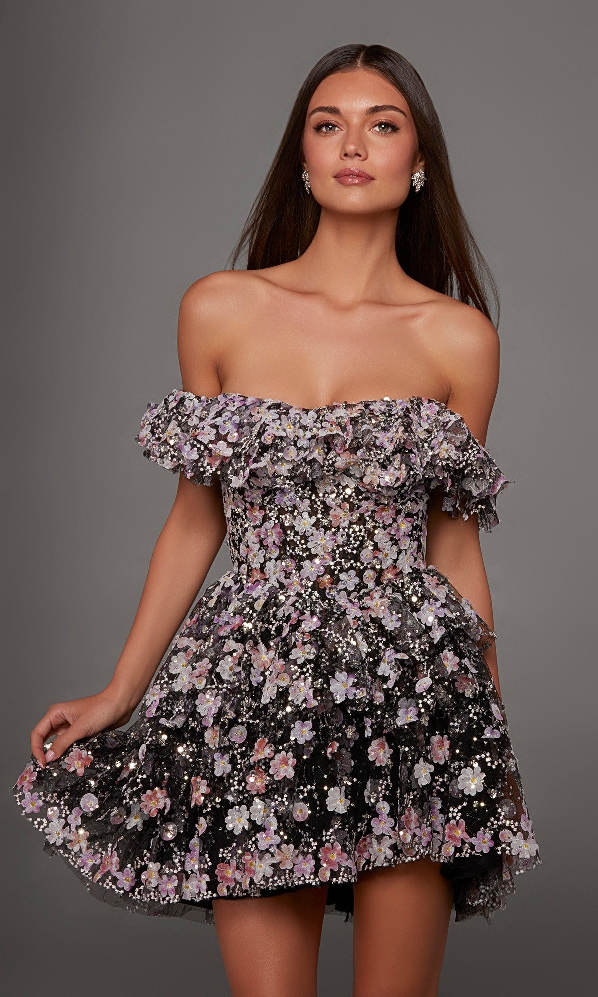 A woman wearing the Alyce 30115 Short Homecoming Dress, featuring an off-shoulder, ruffled design with pink, purple, and white floral patterns, stands against a plain gray background.
