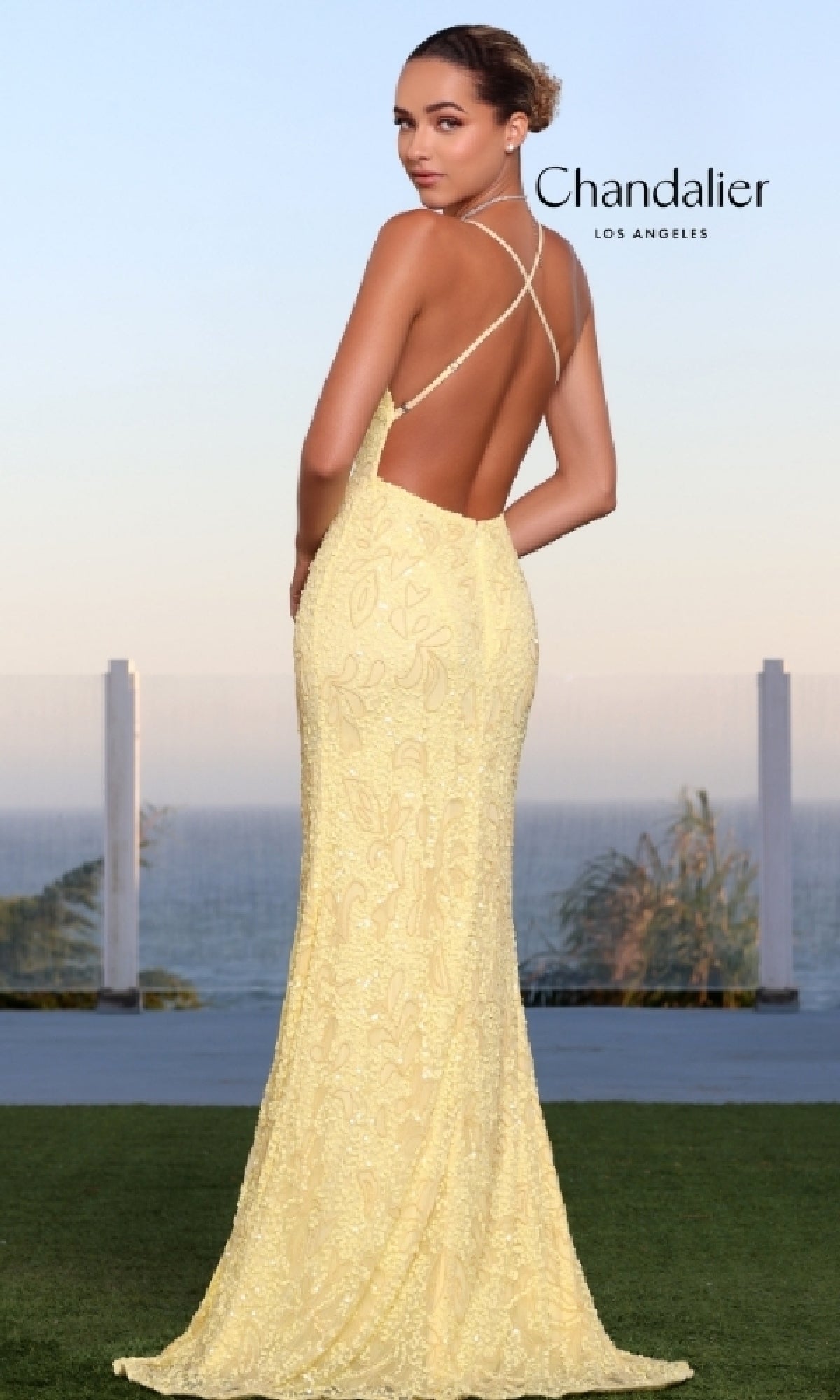 A woman in a yellow Long Prom Dress 30118 by Chandalier stands on grass, facing away, with the ocean and sky behind her. 