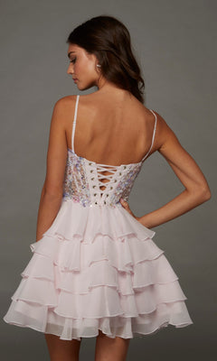 A woman poses with one hand on her hip against a plain background, wearing the Short Homecoming Dress: Alyce 30121—a light pink, fit-and-flare dress featuring a sleeveless, ruffled skirt and a sequin-beaded embellished bodice with a lace-up back.