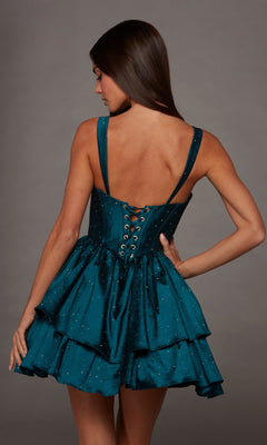 A woman faces away, modeling the Alyce 30122 short homecoming dress. The teal fit-and-flare design features a sleeveless silhouette, lace-up corset back, and subtle rhinestone embellishments, all set against a plain gray background.