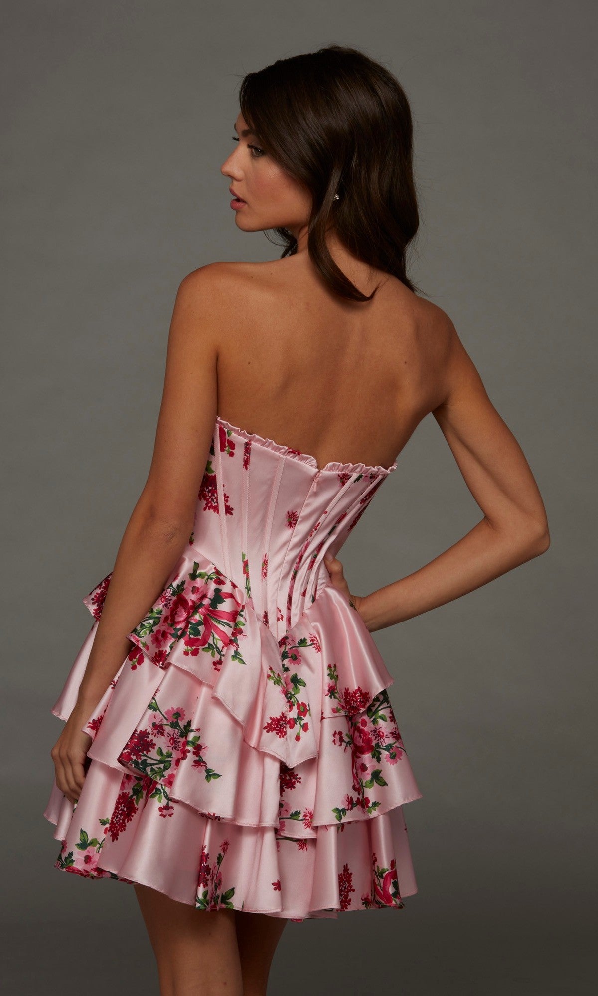 Wearing the Alyce 30123 Short Homecoming Dress—an elegant pink, strapless mini dress featuring floral details, tiered layers, and an A-line silhouette—a woman stands sideways against a plain gray background.