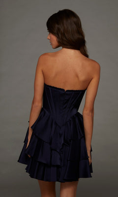 A woman models the Alyce 30123 short homecoming dress—a strapless satin gown featuring a dark blue, ruffled design and a subtle A-line silhouette—as she stands facing away with her head turned slightly to the side against a plain gray background.