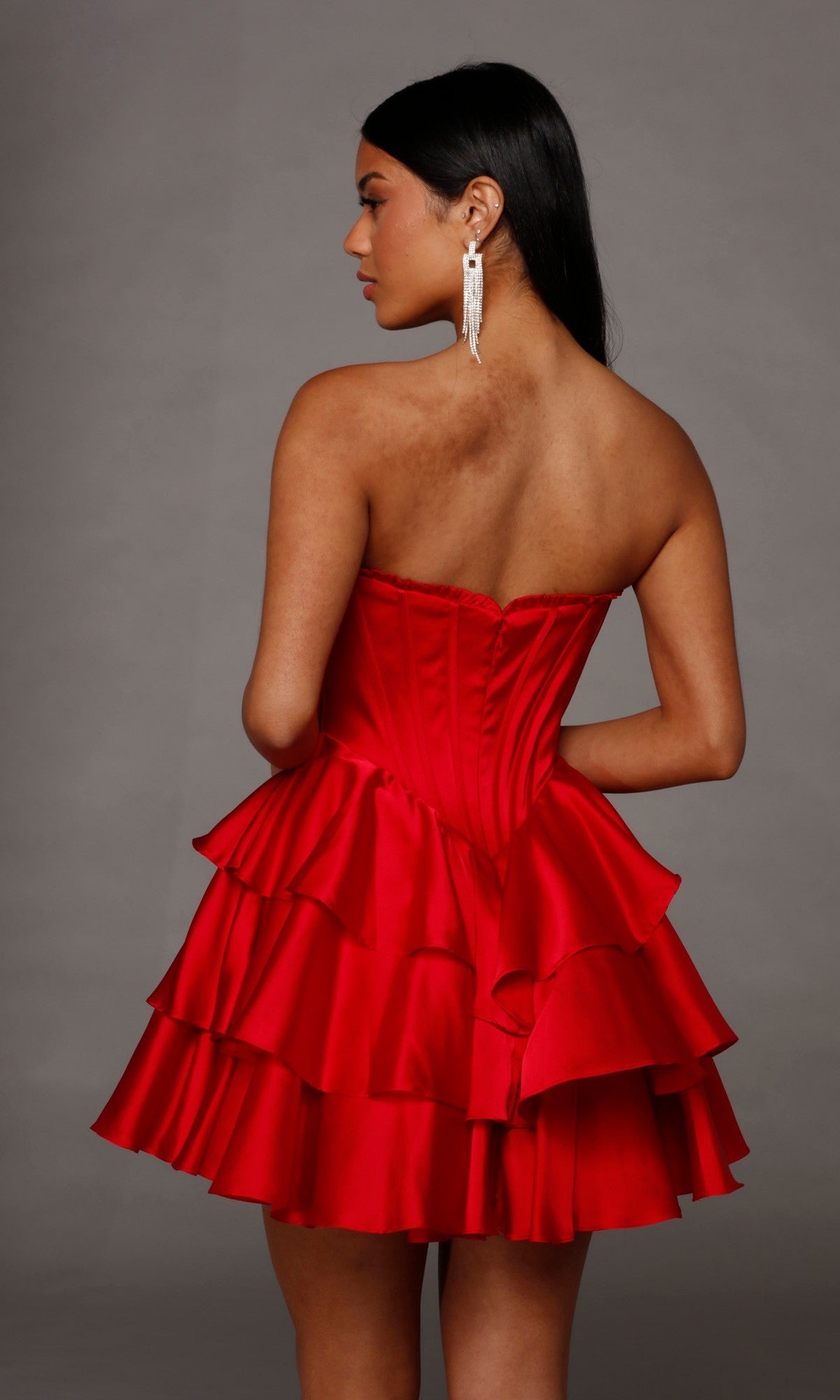 A woman stands with her back to the camera, wearing the Alyce 30123 Short Homecoming Dress—a strapless satin design with a ruffled skirt, elegant corset bodice, and graceful A-line silhouette. She pairs the dress with long silver earrings while posing against a plain gray background.