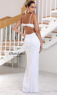 A woman wearing the Long Prom Dress 30123 by Chandalier— a fitted white formal gown with thin straps and an open back—stands on a marble floor before a staircase. The "Chandalier Los Angeles" logo appears on the image’s left side.