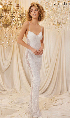 A woman wearing the Long Prom Dress 30124 by Chandalier—a fitted white sequin gown with thin straps—stands before draped fabric and ornate chandeliers.