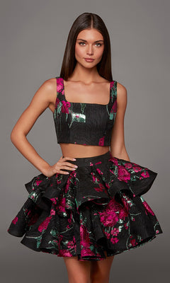 A woman stands against a plain gray background wearing the Alyce 30126 Short Homecoming Dress, a stylish two-piece ensemble in black and fuchsia with a square-neck crop top and a ruffled skirt adorned with pink and green floral patterns.