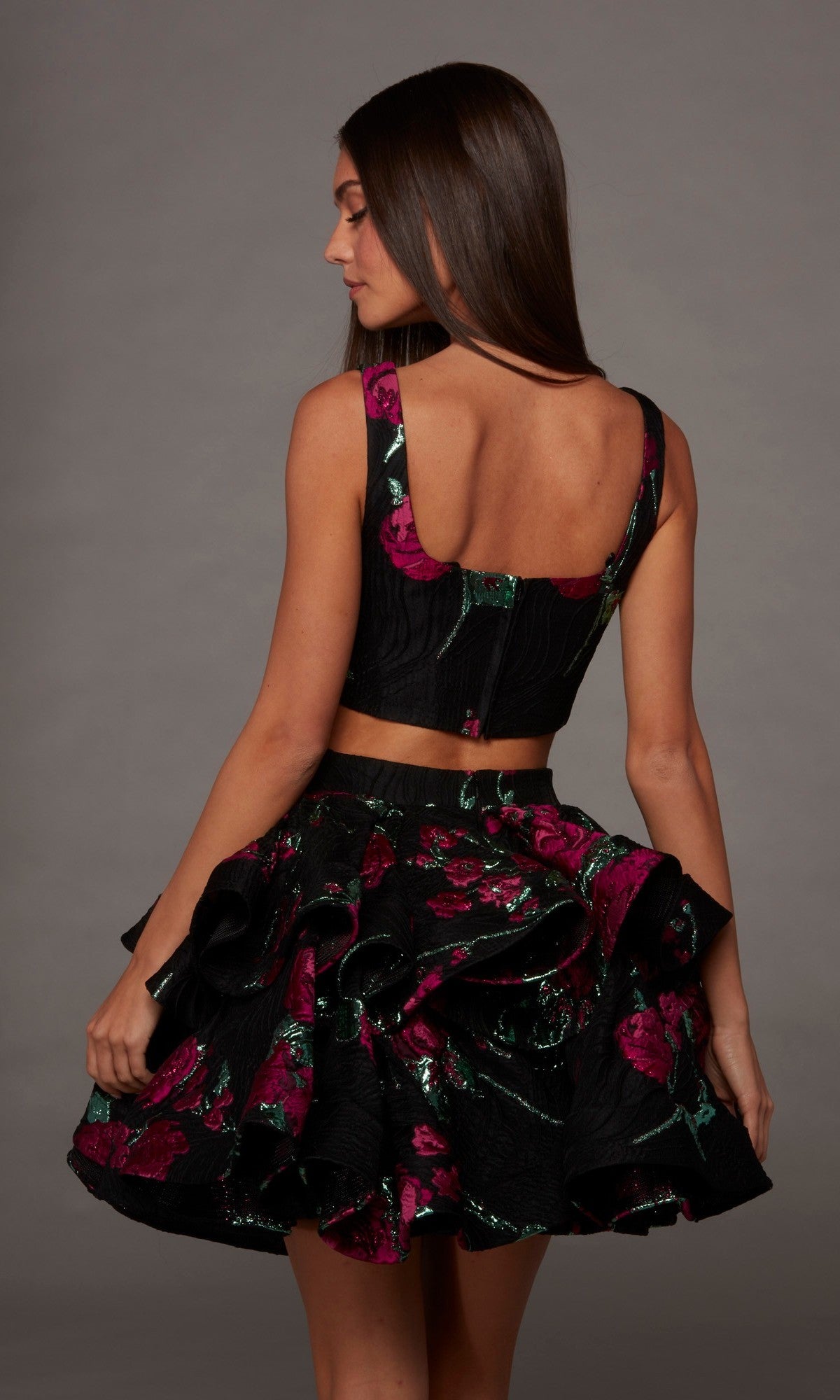 Wearing the Alyce 30126 short homecoming dress, a woman stands against a plain gray background; the sleeveless black crop top features a square neckline and is paired with a ruffled floral skirt adorned with pink and green patterns.