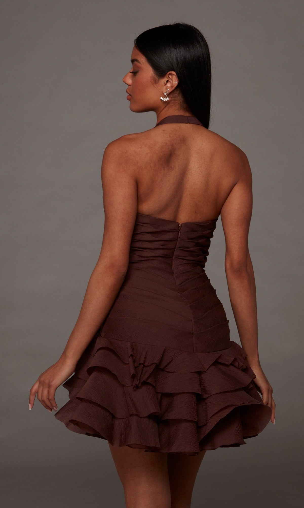 A woman stands facing away against a plain gray background, wearing the Alyce 30127 Short Homecoming Dress—a strapless, brown ruffled crinkle organza design.