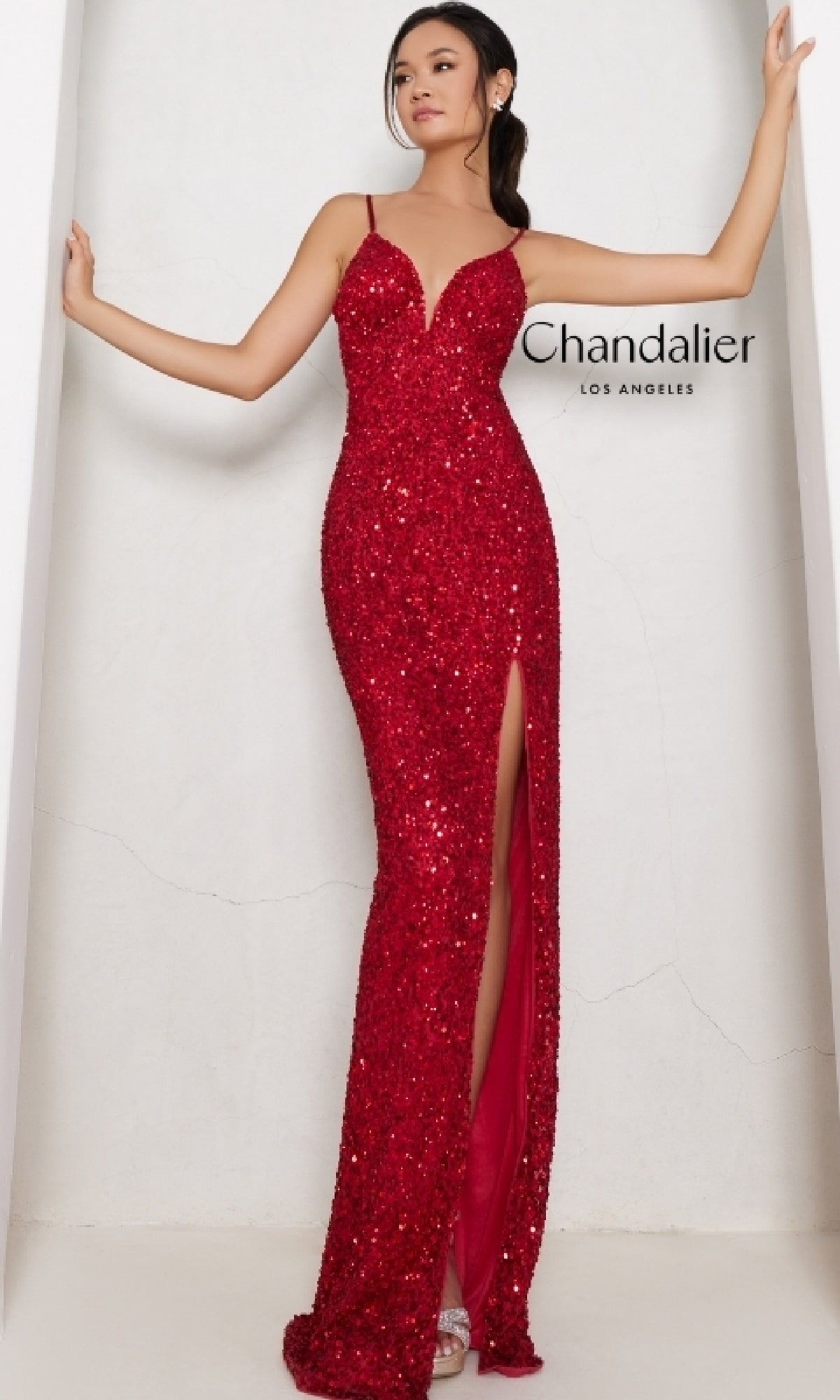 A woman poses against a white wall, wearing the sleeveless red sequin Long Prom Dress: Chandalier 30179 with a thigh-high slit. The text 