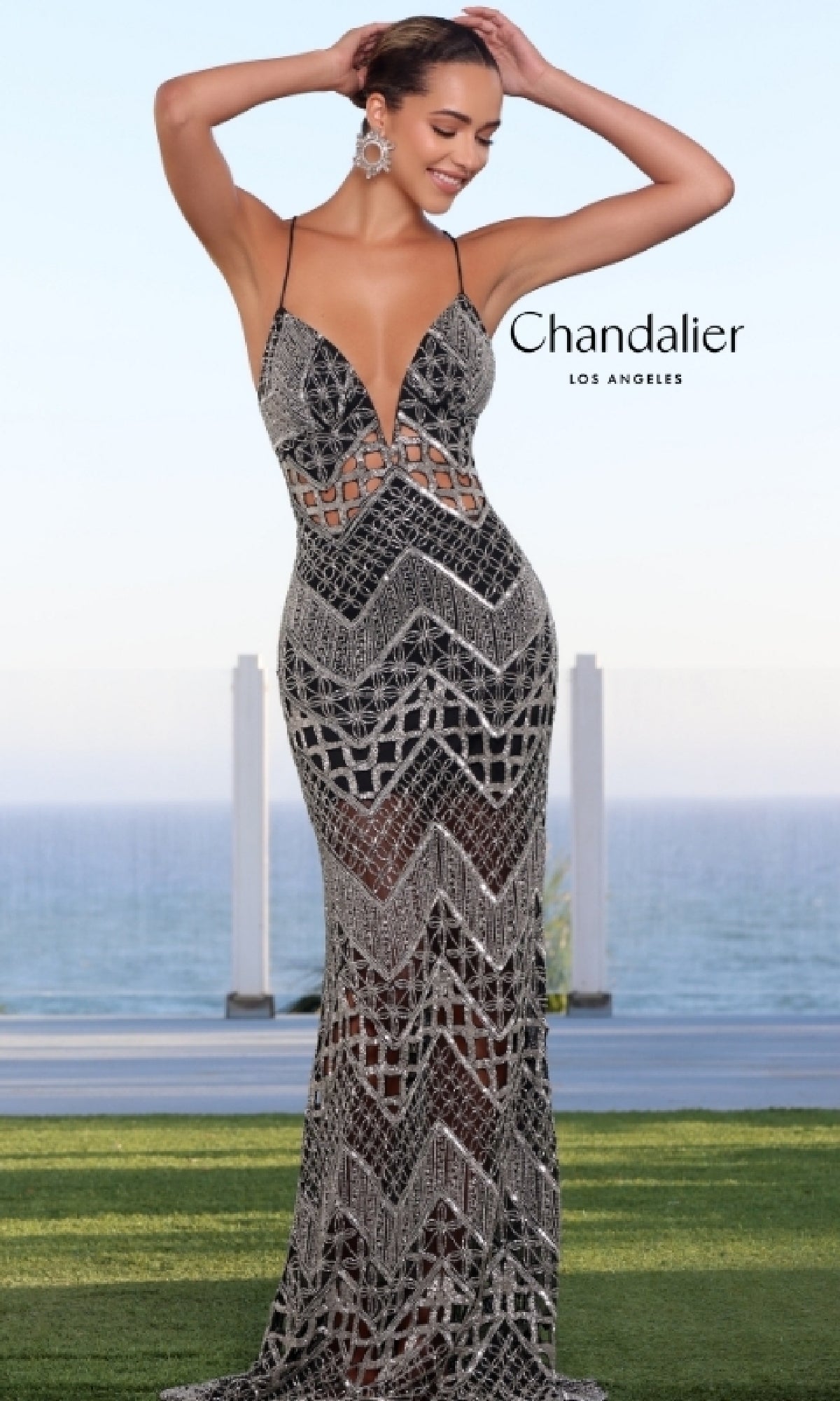 A woman poses outdoors by the ocean and sky in the Chandalier 30181 Long Prom Dress, featuring a sparkly silver and black geometric pattern, thin straps, and an open back. 