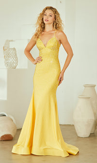 A woman wears the Open-Back Sparkly Long Beaded Prom Dress 3018, a fitted, floor-length yellow gown with thin straps, as she stands indoors near white decorative pottery.