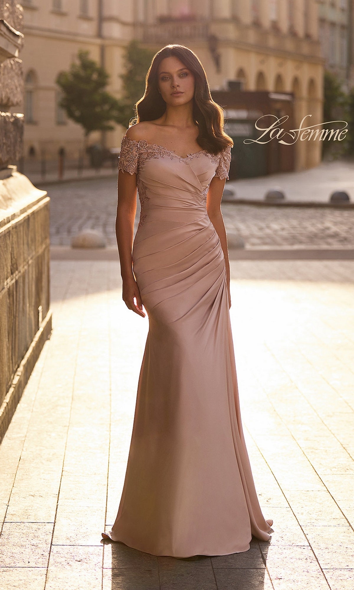 A woman stands on a cobblestone street wearing a champagne-colored La Femme 30199 long formal dress, featuring an off-the-shoulder design with lace detailing on the sleeves and neckline.