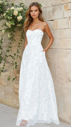 A woman models the Blondie Nites 3024BN, a long off-white strapless formal dress with a subtle light blue floral pattern, standing by a stone wall adorned with green foliage.