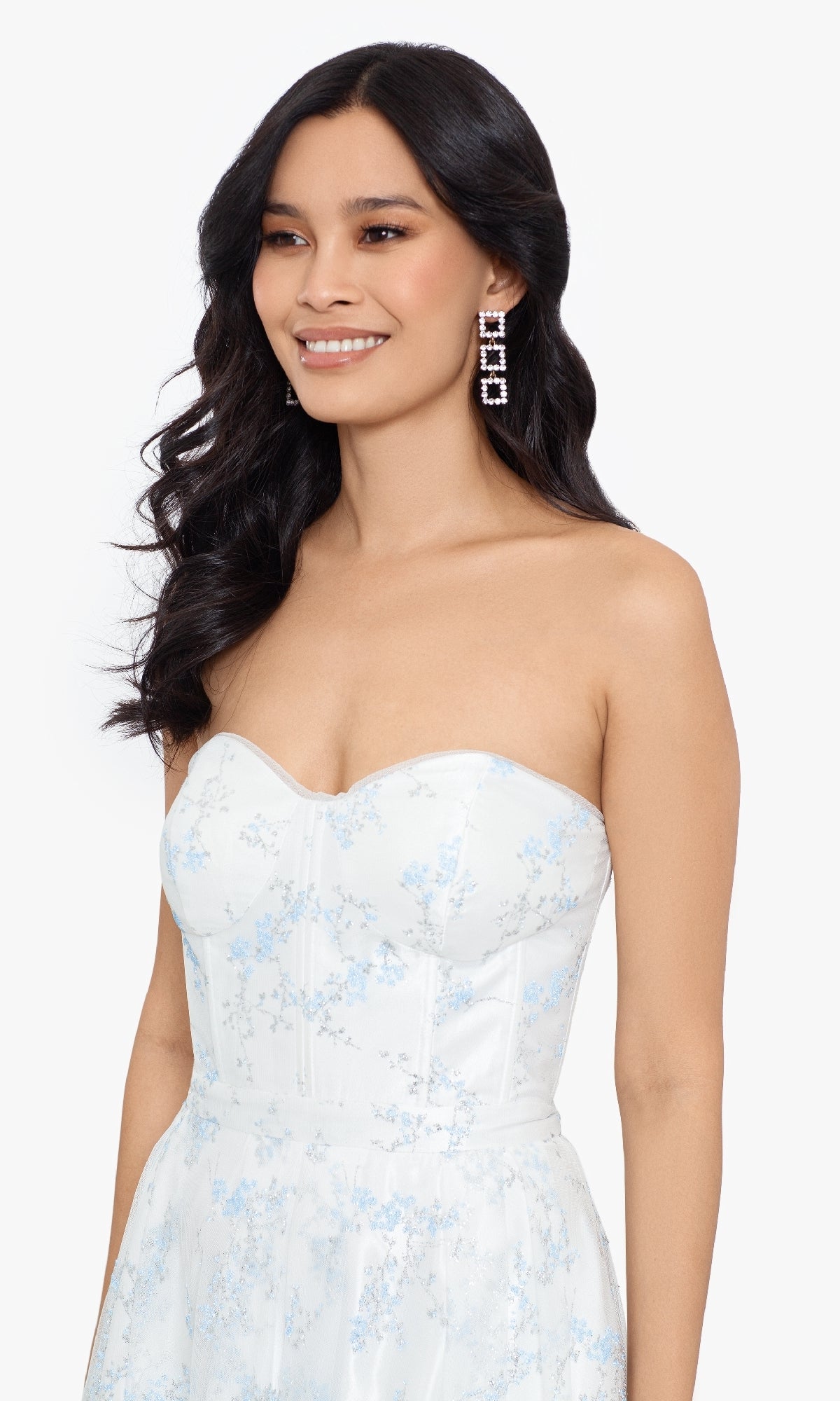 A woman with long wavy hair and large geometric earrings wears the Blondie Nites 3024BN long formal dress—an off-white, strapless gown with light blue floral patterns—standing against a plain background.