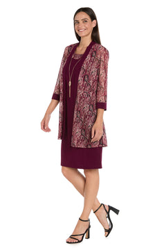 A woman stands against a white background wearing the Petite Short Wine Party Dress: R&M Richards 3029P, paired with a patterned sheer jacket, black heeled sandals, and a long necklace.