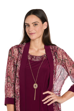 A woman with long brown hair confidently poses in the R&M Richards 3029P Petite Short Wine Party Dress, perfect for semi-formal occasions. She accessorizes with a gold pendant necklace over a maroon top.
