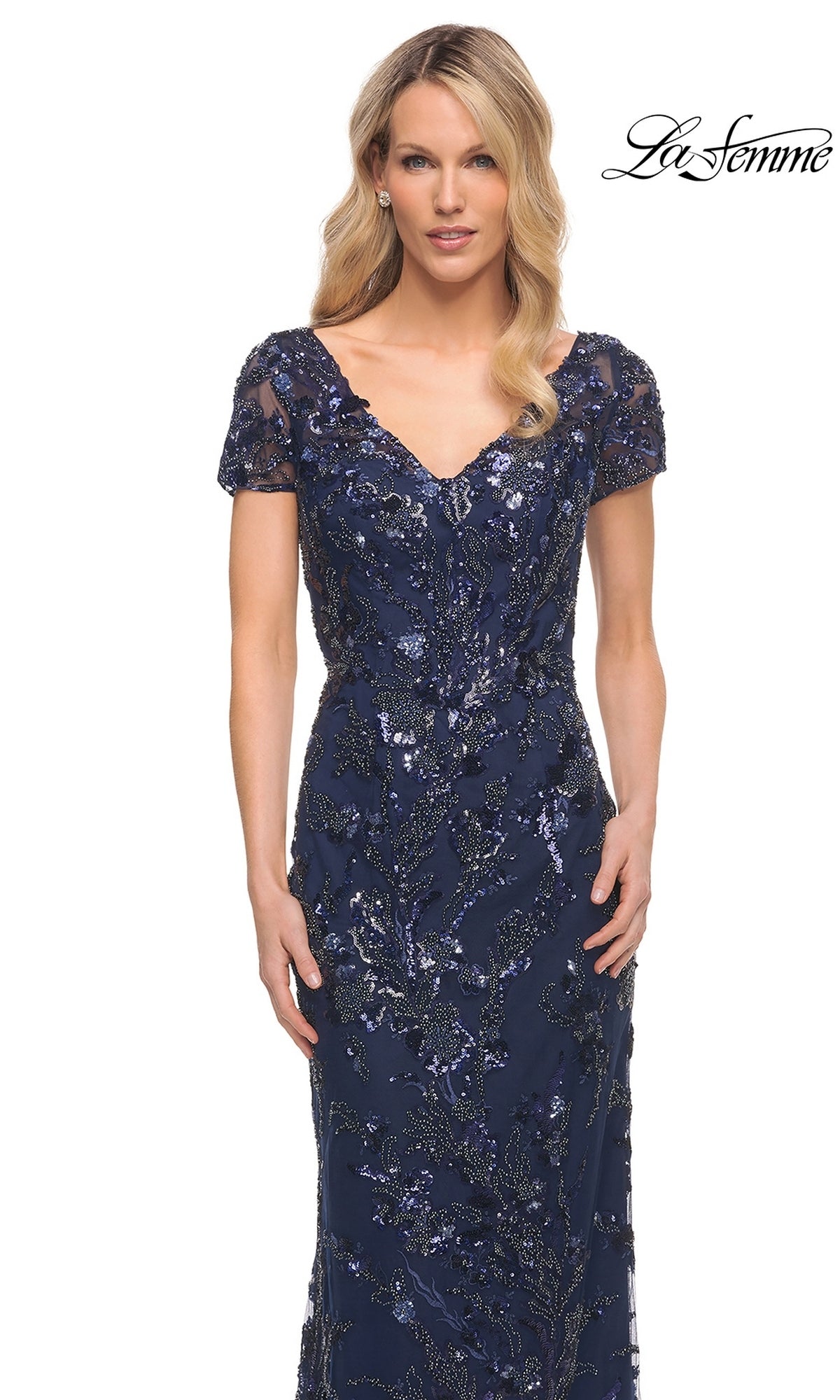 A woman wears the Long Formal Dress: La Femme 30375, a navy blue, short-sleeved beaded gown with a V-neckline and intricate leafy sequin embellishments.