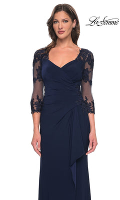 Woman in Long Formal Dress: La Femme 30384—navy blue jersey with sheer lace sleeves and floral embroidery, standing against a white background. “La Femme” logo top right.