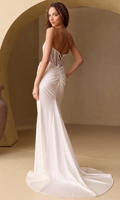 A woman models the Amelia Couture 3042 Beaded-Bodice Long Prom Dress, featuring a strapless white fitted bodice, lace-up back, and a ruched train, set against a neutral arched background.