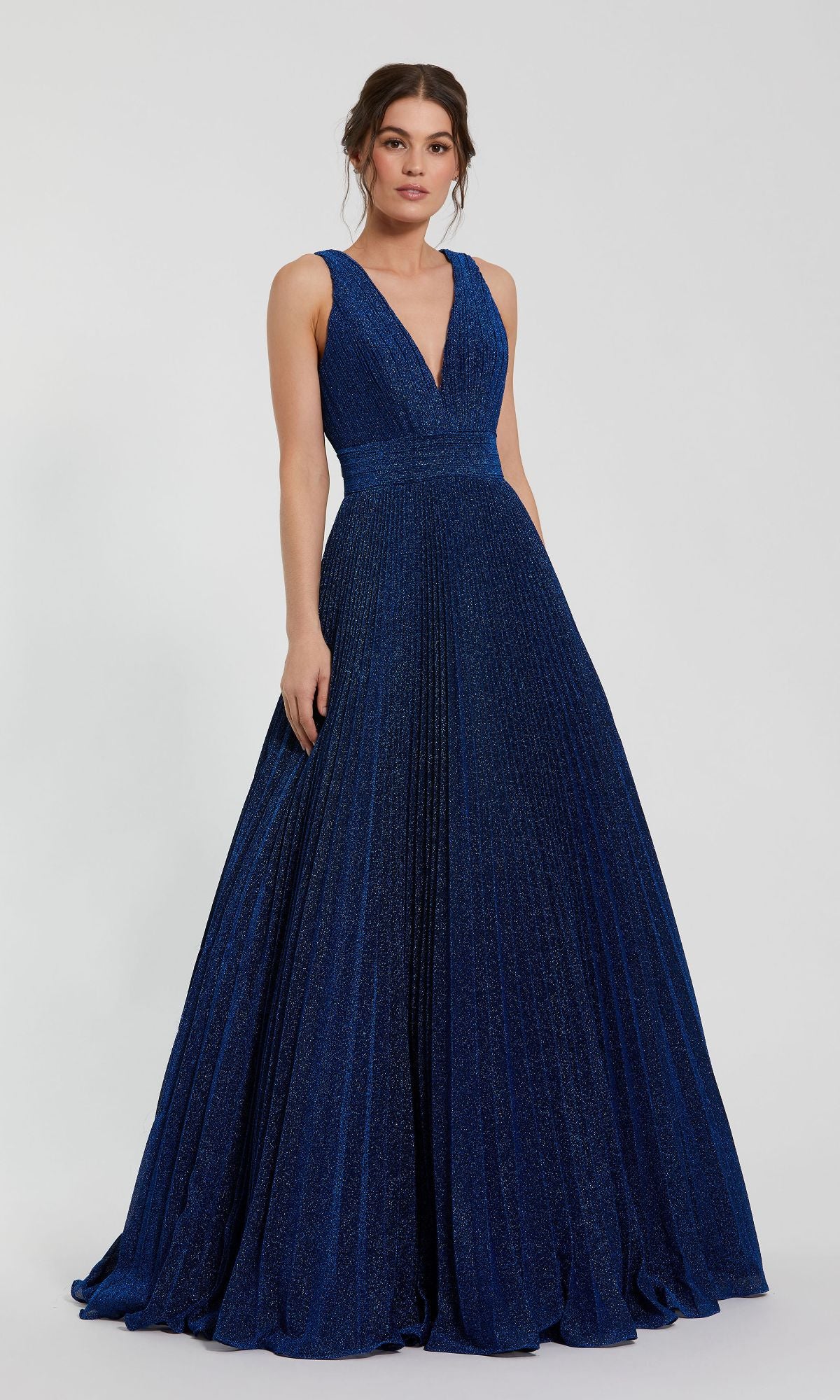 A woman in the Mac Duggal 30773 Long Pleated A-Line Prom Dress, featuring a sleeveless deep V-neck and flowing blue skirt, stands against a plain light background.