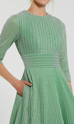 A woman wears the Sage Long Pleated Formal Dress by Mac Duggal (30775) in green, featuring shimmery pleats, an A-line silhouette, three-quarter sleeves, and a side pocket where she tucks her right hand.