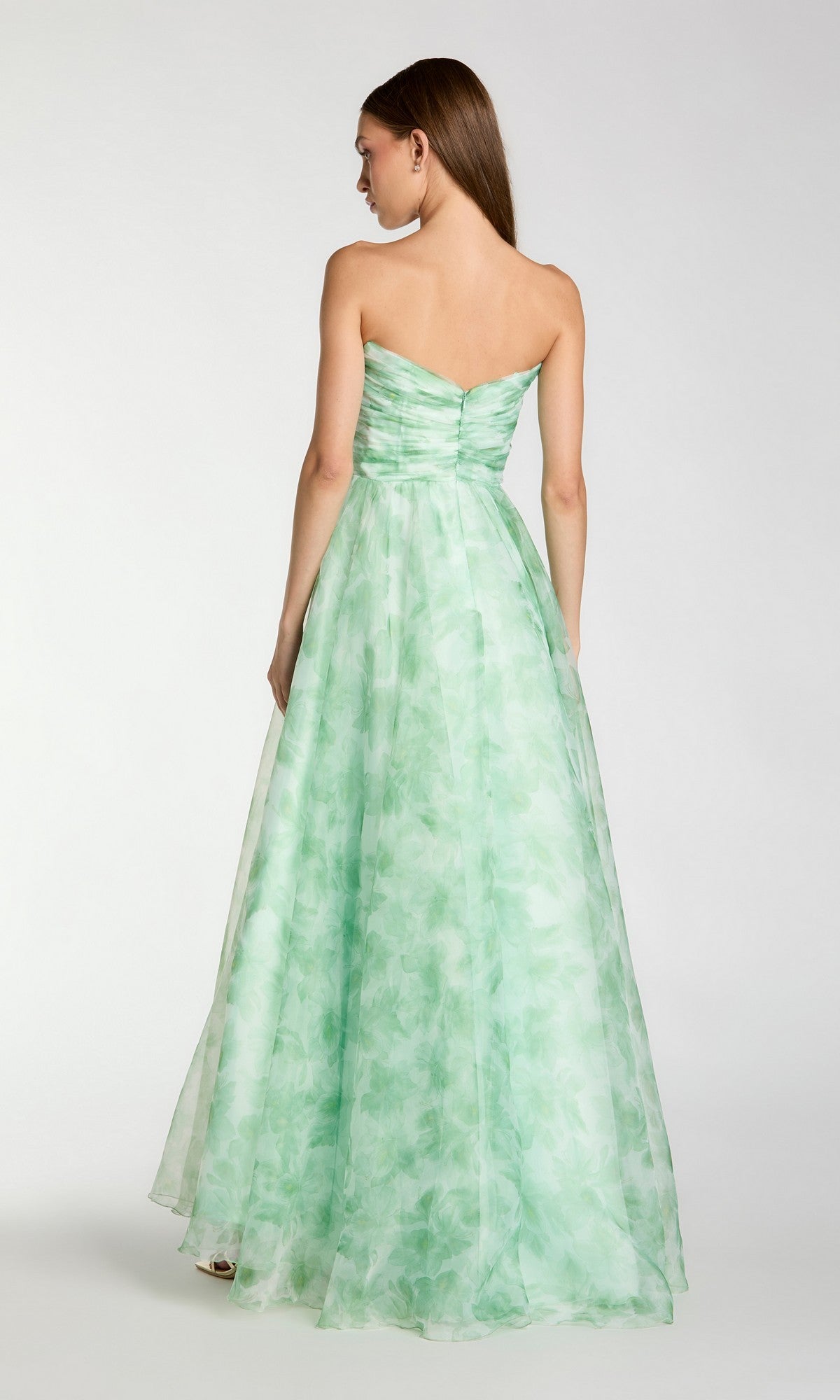 Woman in a Mac Duggal 30783 long formal dress, featuring a strapless ruched bodice and subtle floral pattern in light green, standing floor-length and facing away from the camera.