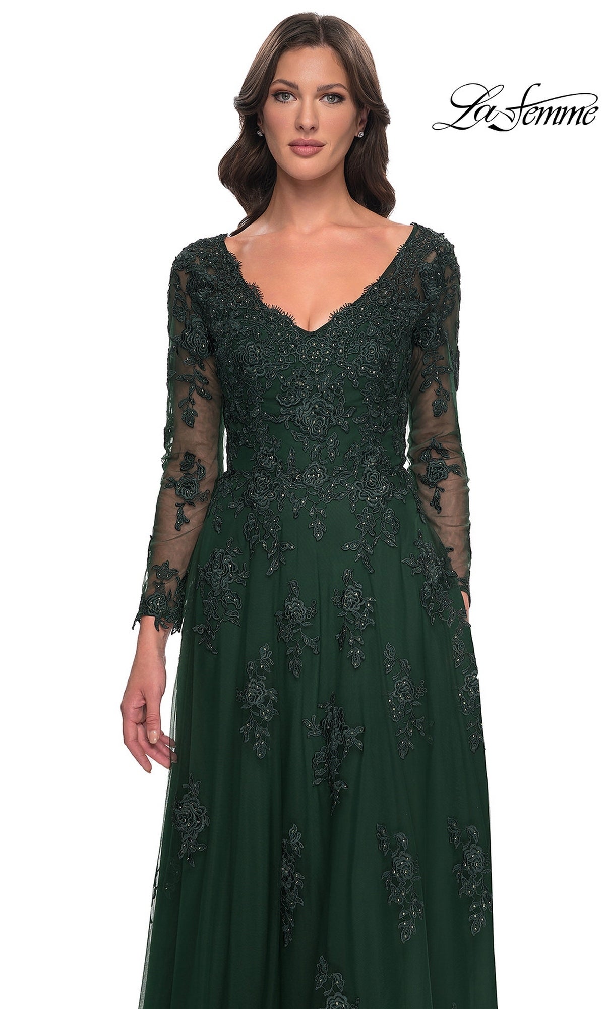 A woman wears the Long Formal Dress: La Femme 30795, featuring a dark green gown with long sleeves, a V-neckline, and floral lace embroidery atop a tulle A-line skirt.