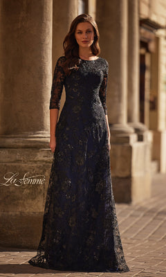 A woman in the Black and Blue Lace Formal Gown: La Femme 30825, featuring three-quarter sleeves, stands outdoors before stone columns.