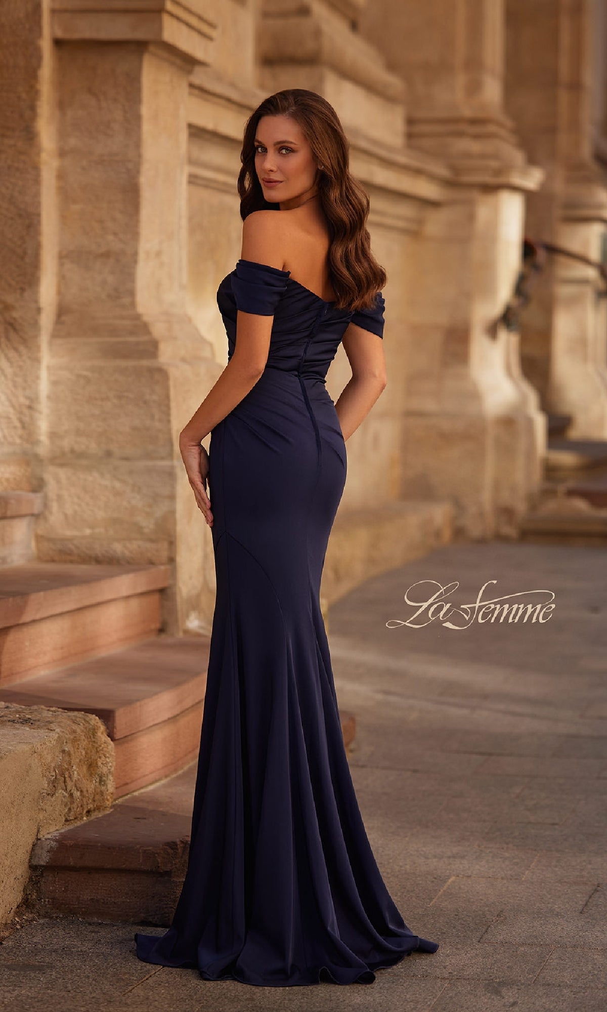 A woman stands outdoors on stone steps in the Off-the-Shoulder Long Formal Dress: La Femme 30846, a fitted mermaid gown with a flowing skirt that creates an elegant silhouette as she looks over her shoulder.