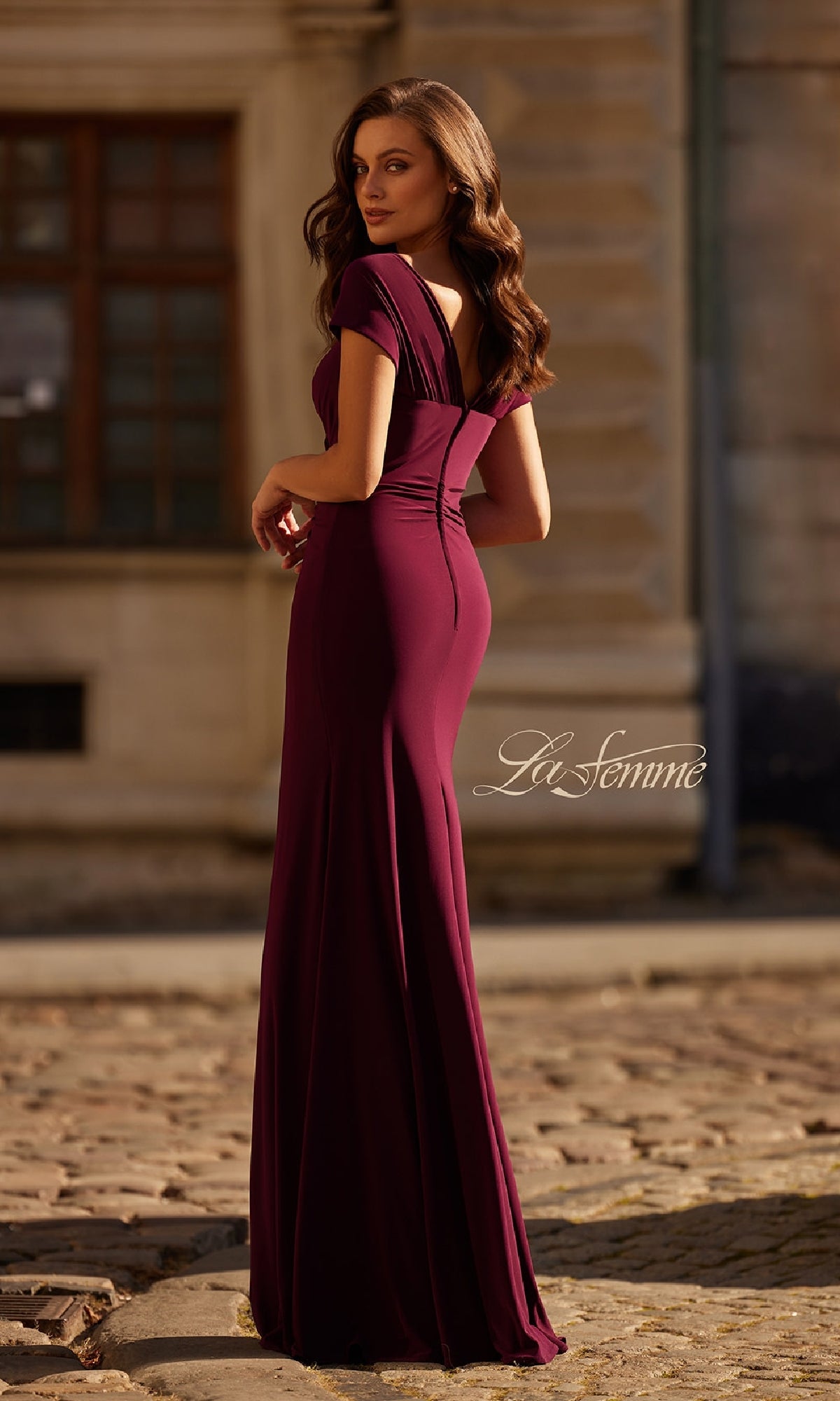 A woman stands outdoors on a cobblestone street, looking back over her shoulder in the La Femme 30874 Cap-Sleeve Long Jersey Formal Dress. The deep burgundy gown features a fitted silhouette and elegant details.