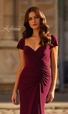 A woman stands outdoors before a stone building, wearing the La Femme 30874 cap-sleeve long jersey formal dress in deep burgundy with ruched details.