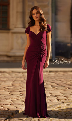 A woman in the Cap-Sleeve Long Jersey Formal Dress: La Femme 30874, featuring a fitted, floor-length burgundy design with ruched details, stands on a cobblestone street before a stone building.