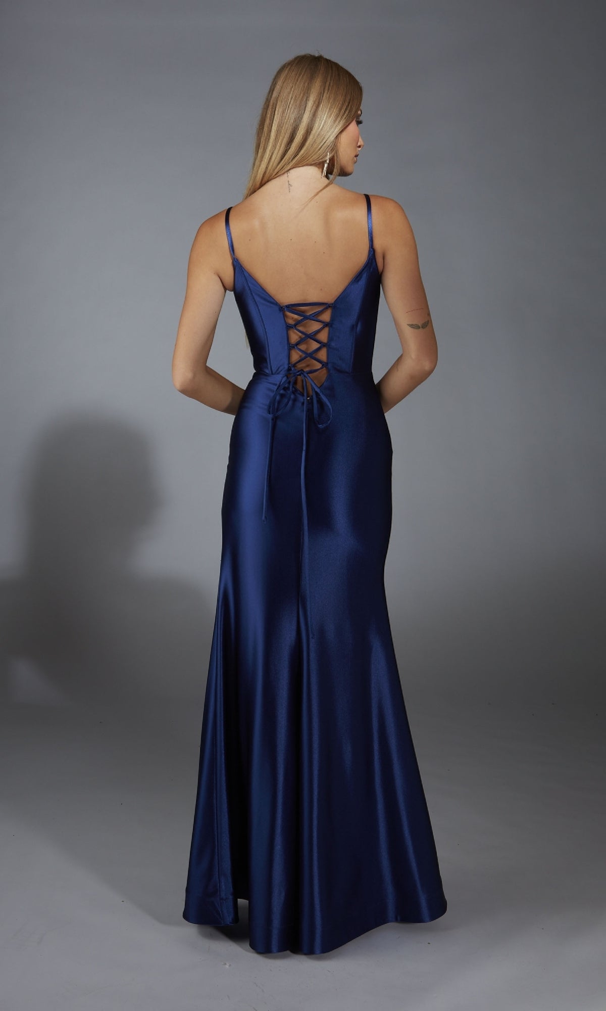 A woman in the Long Navy Blue Prom Dress: Aerie Couture 309, featuring a corset bodice, lace-up back, thin straps, and a floor-length satin skirt, stands against a plain gray background.