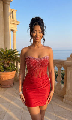 A woman stands on a sunlit balcony framed by columns and a potted palm, overlooking the sparkling sea, wearing the Amelia Couture 3108S red strapless mini homecoming dress with a sheer corset bodice.