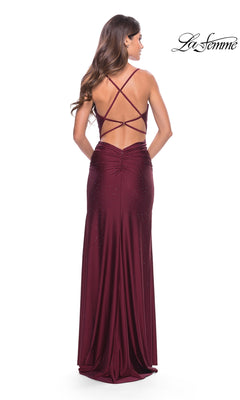 A woman wears the La Femme Cowl-Neck Long Beaded Prom Dress 31123: a backless, floor-length burgundy gown with crisscross straps, delicate sparkling embellishments, and a hint of cowl neckline. "La Femme" logo appears in the top right corner.