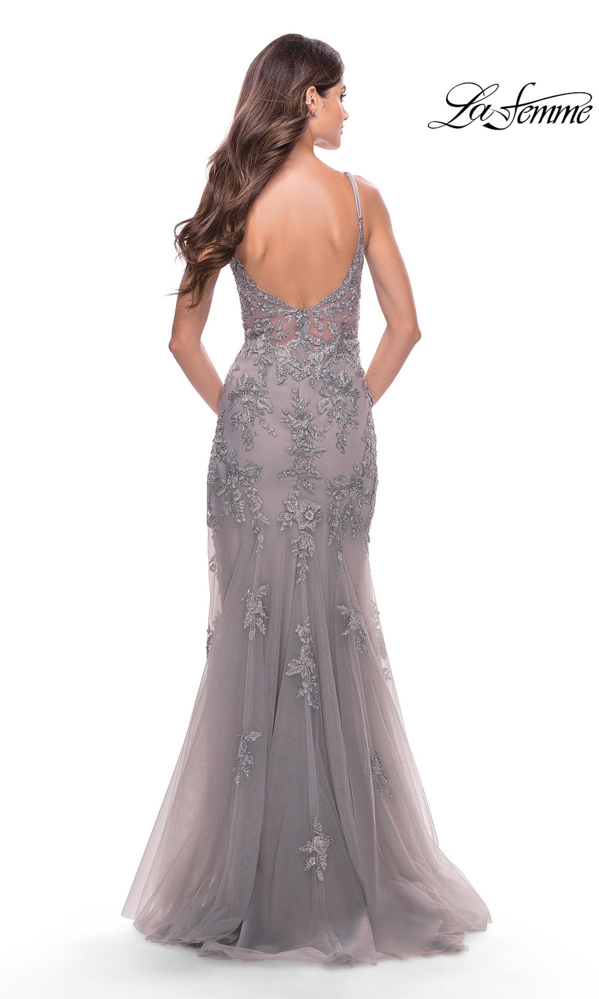 A woman with long brown hair wears the La Femme Long Lace Prom Dress with a sheer bodice, featuring gray floral lace and a low back, as she stands against a white background.