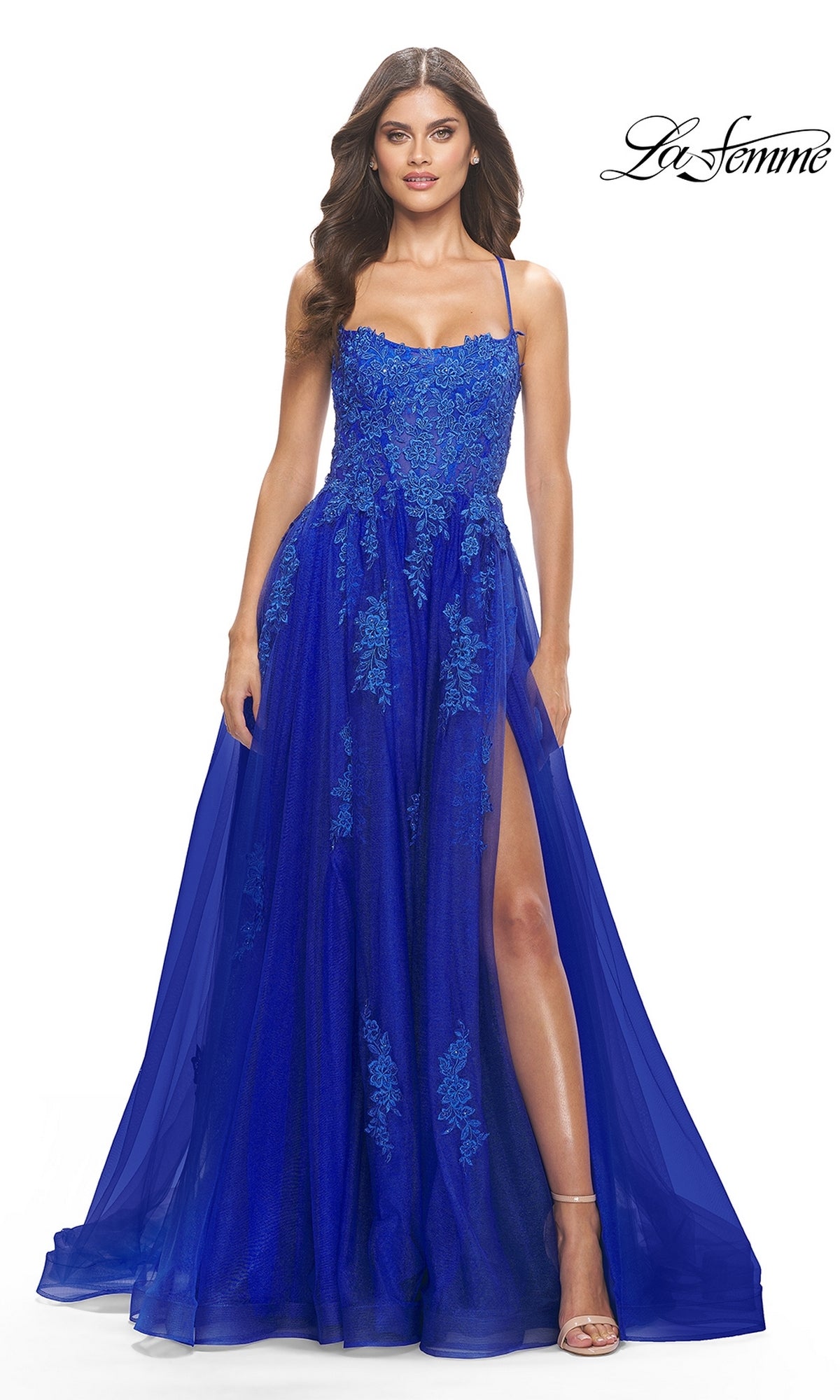 A woman in the La Femme Long A-Line Lace Prom Ball Gown 31135, featuring royal blue floral lace, thin straps, a sheer overlay, and a thigh-high slit, stands against a white background.