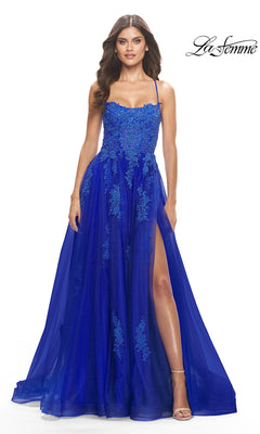 A woman in the La Femme Long A-Line Lace Prom Ball Gown 31135, featuring royal blue floral lace, thin straps, a sheer overlay, and a thigh-high slit, stands against a white background.