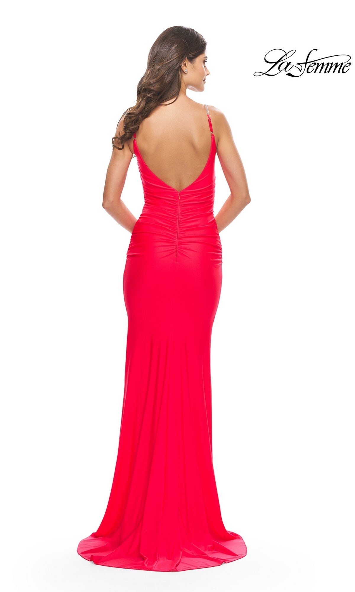 A woman stands facing away from the camera in the La Femme Ruched Fitted Designer Prom Dress 31222, a bright pink, floor-length gown with a v-neck and deep V-back. The La Femme logo is in the top right corner.