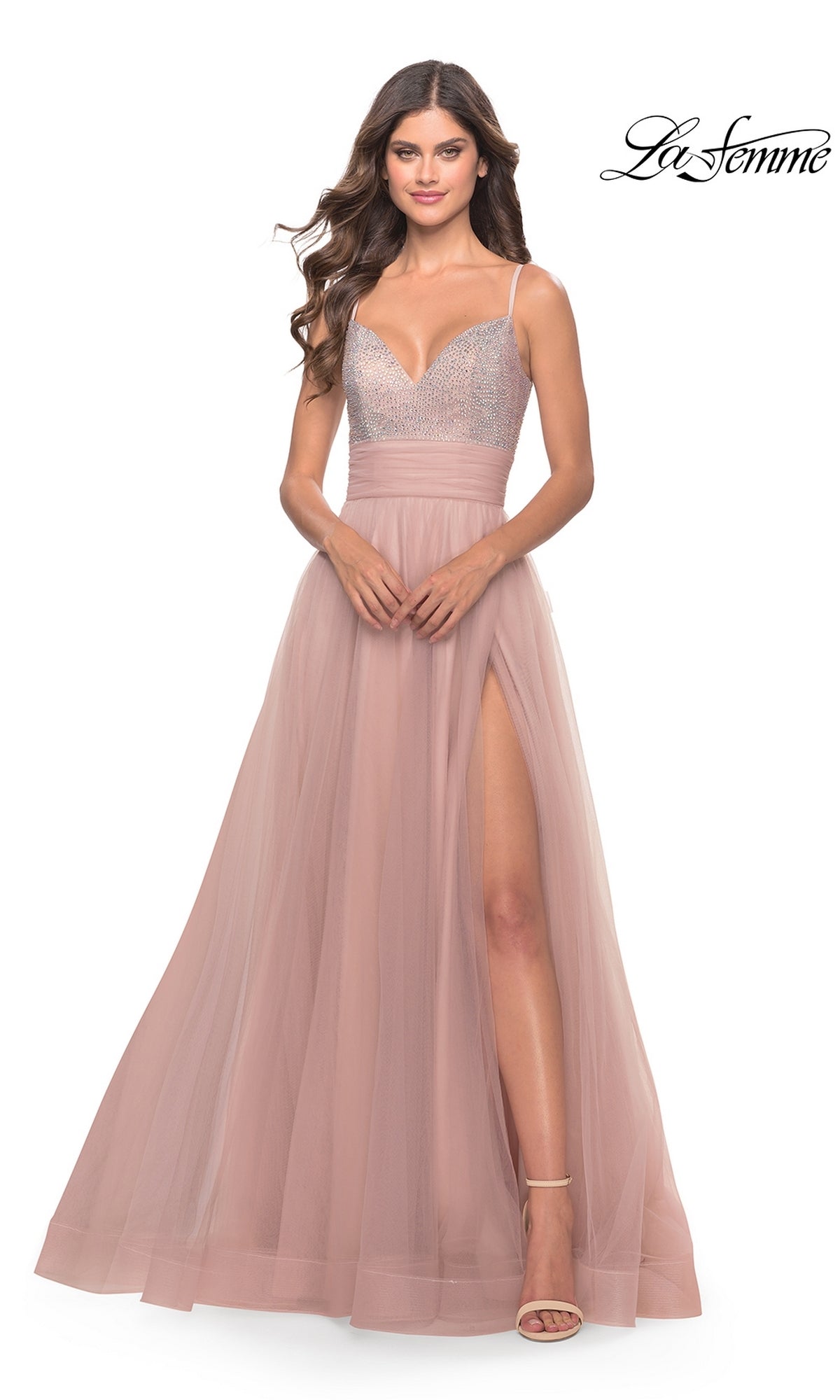 A woman wears the Beaded-Bodice Tulle Prom Ball Gown 31238, a sleeveless blush pink dress with a v-neck, beaded bodice, tulle skirt, and high slit, standing against a white background. 