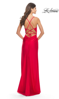 Woman in the La Femme Fringe-Slit Long Prom Dress 31326, a bright red gown with crisscross straps, tie-back detail, and beaded fringe, stands facing away; "La Femme" logo appears in the upper right corner.