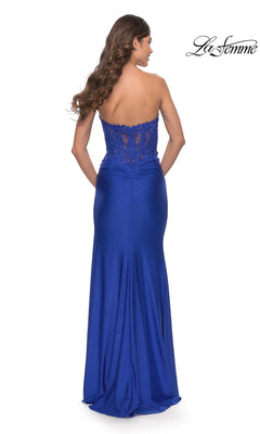 A woman showcases the elegant La Femme Strapless Sheer-Waist Prom Dress 31343—a floor-length blue gown with a fitted waist, lace back detail, and glitter accents—standing with her back to the camera.