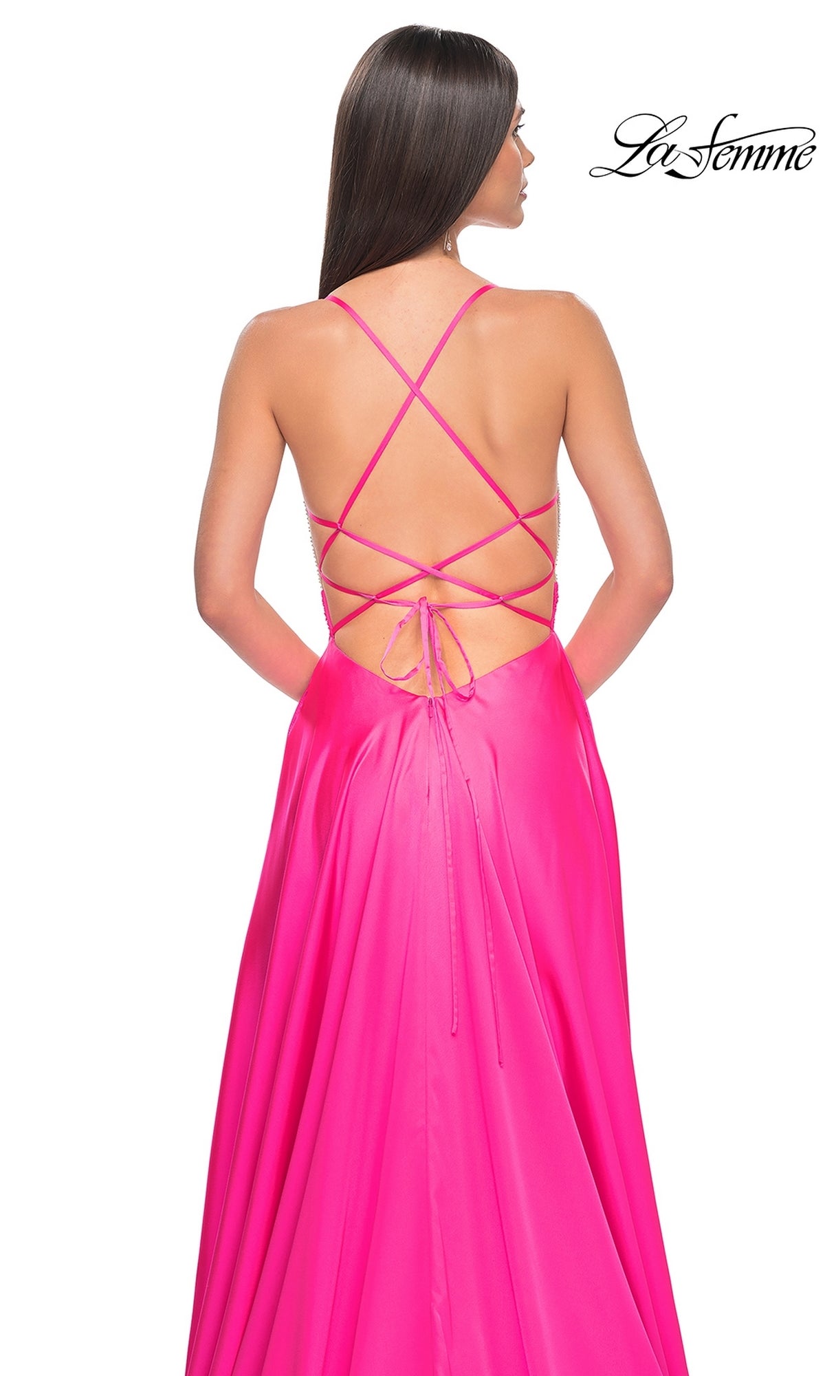 A woman wears the La Femme Neon Pink Long Prom Dress 31448, featuring a beaded bodice, open back, and crisscross lace-up straps, standing against a white background with the 