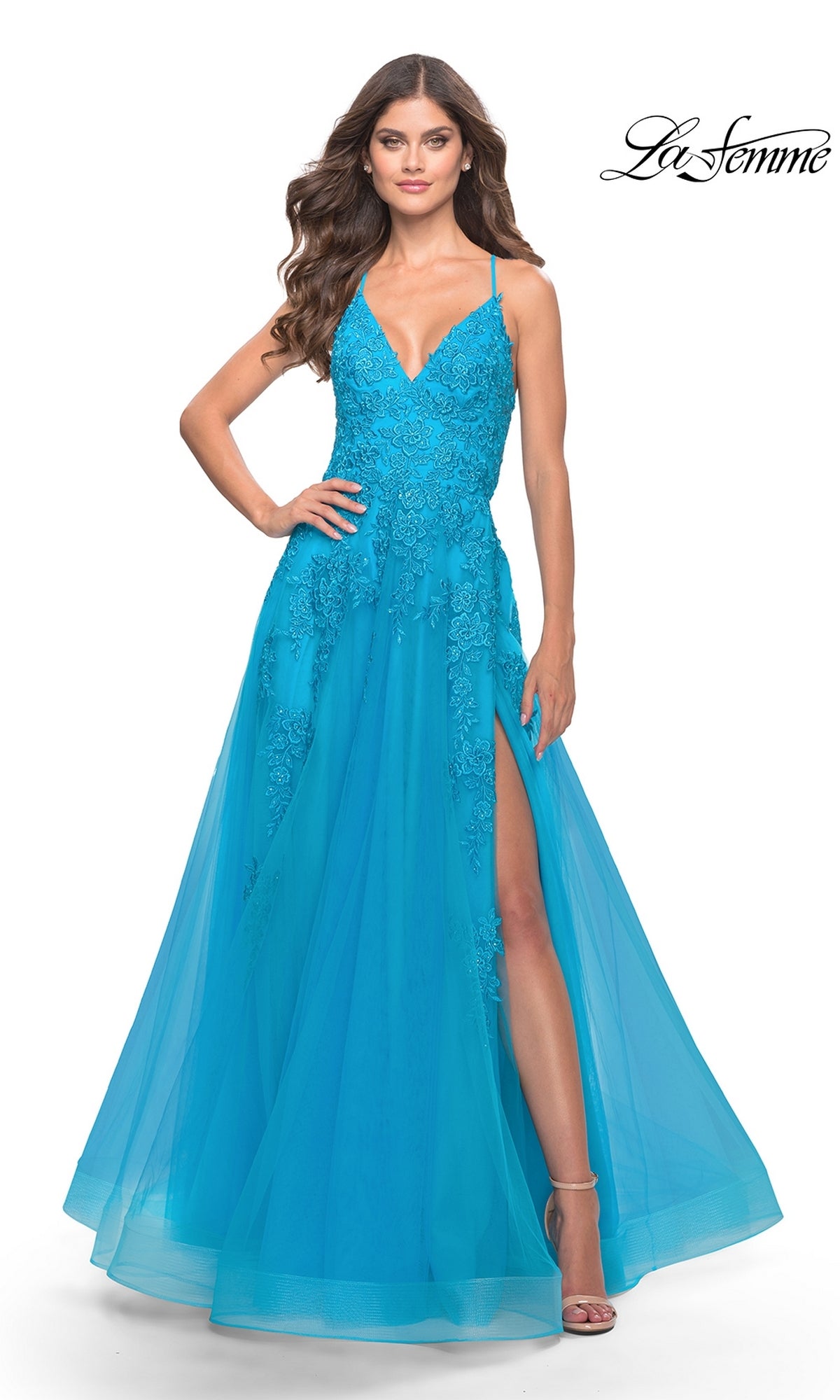 A woman stands with one hand on her hip, wearing the La Femme Strappy-Back Long A-Line Prom Dress 31503 in bright blue with lace detailing, a V-neckline, high slit, and strappy-back design against a white background.