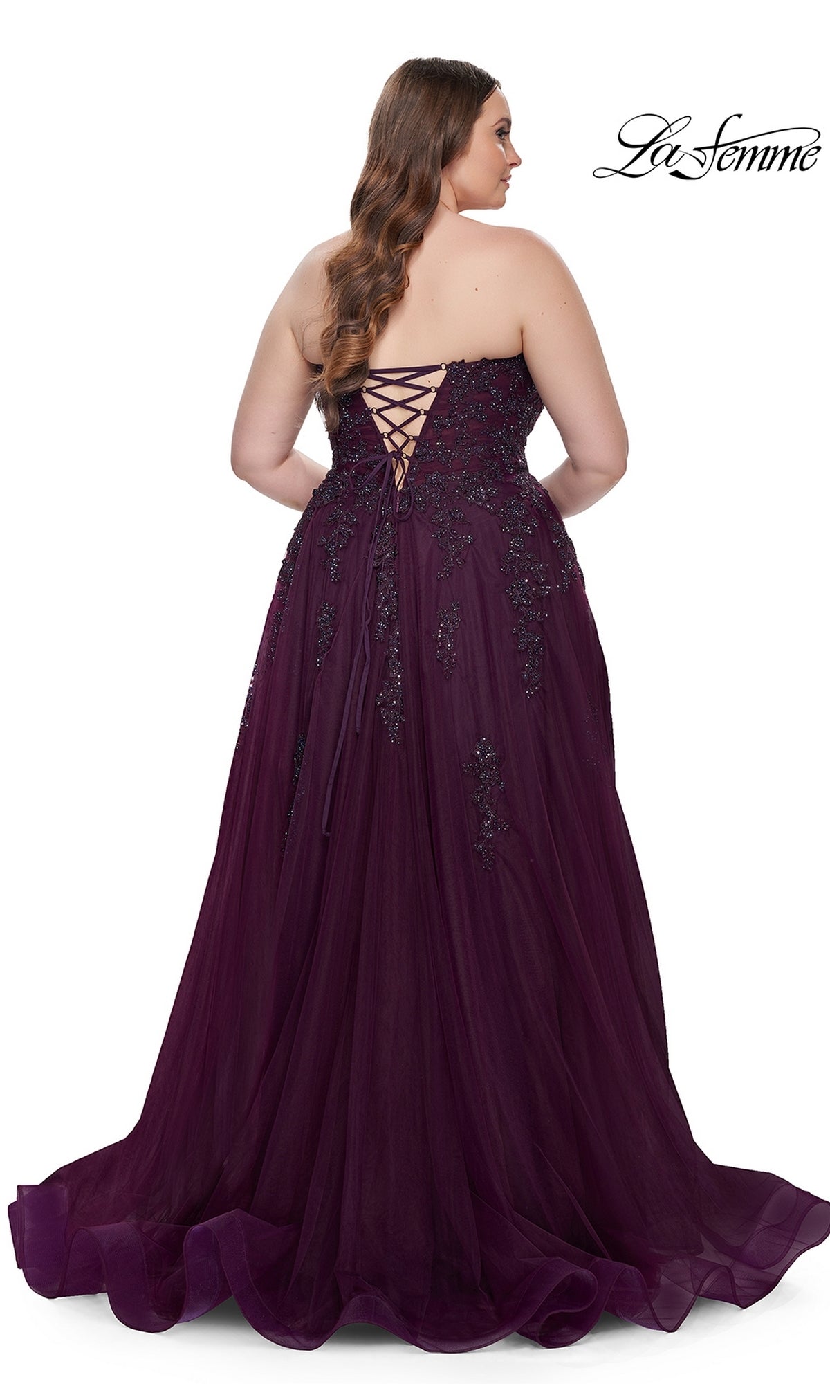 A woman in the La Femme Long Prom Dress 31570, a plus-size strapless purple ball gown with a corset-style bodice and lace-up back, stands facing away from the camera in a floor-length design.