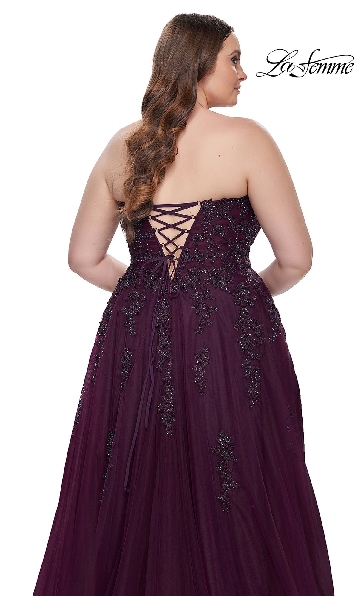 A woman models the La Femme Long Prom Dress 31570, a plus-size strapless gown with a corset-style bodice, beaded embellishments, and an elegant lace-up back as she faces away from the camera.