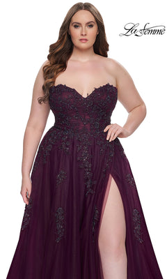 A woman wears the La Femme Long Prom Dress 31570, a plus-size strapless dark purple gown with a corset-style bodice, sweetheart neckline, beaded embellishments, and a thigh-high slit, standing against a white background.