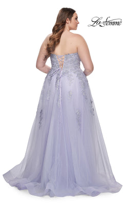 A woman wearing the La Femme Long Prom Dress 31570, a plus-size strapless gown with a corset-style bodice, lace-up back, and embroidered floral lace, stands with her back to the camera.