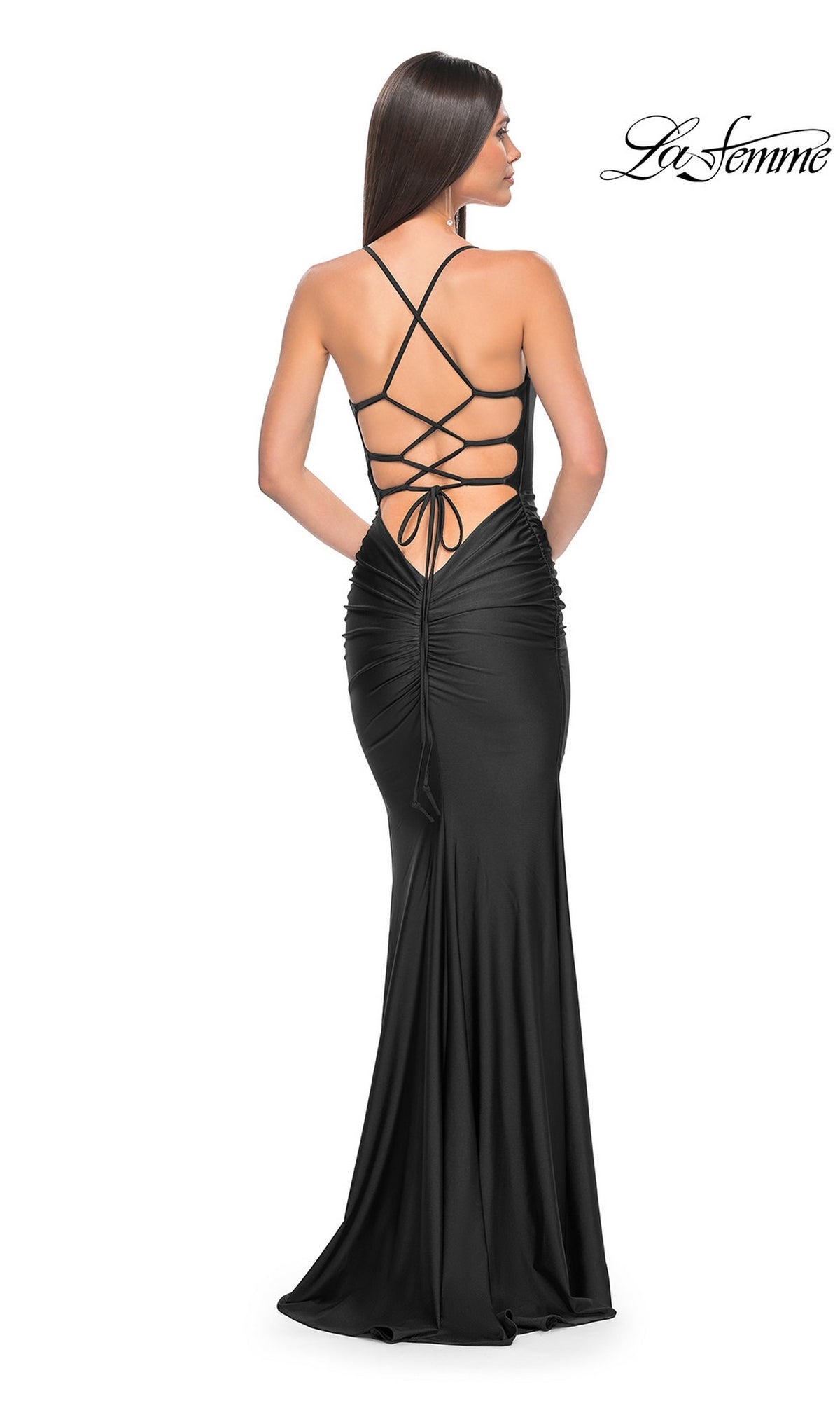 A woman in the La Femme Strappy-Back Simple Long Prom Dress 31618, a fitted black gown with an open back and crisscross straps, stands with her hands behind her.