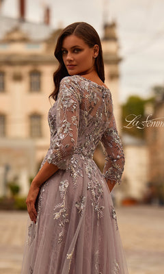 A woman stands outdoors wearing the La Femme 31717 Long Sleeve Mauve Formal Gown, featuring silver floral embroidery and sheer sleeves, as she looks over her shoulder.